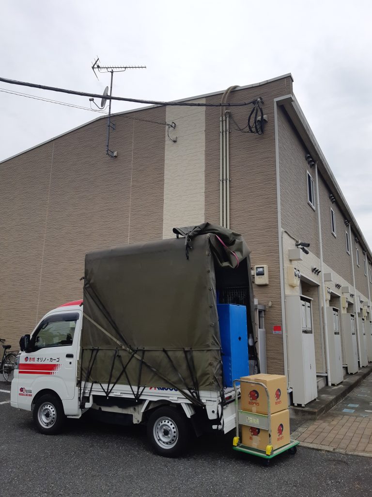 東京都墨田区から群馬県高崎市への単身引越し。引越先のアパートで荷下ろし作業中の赤帽車。台車を使用して、ダンボールとハンガーボックスを運びます。